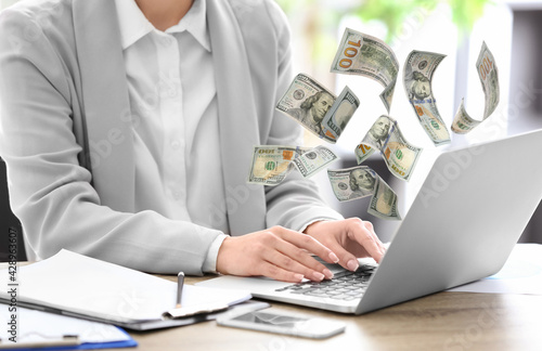 Making money online. Closeup view of woman using laptop at table and flying dollars
