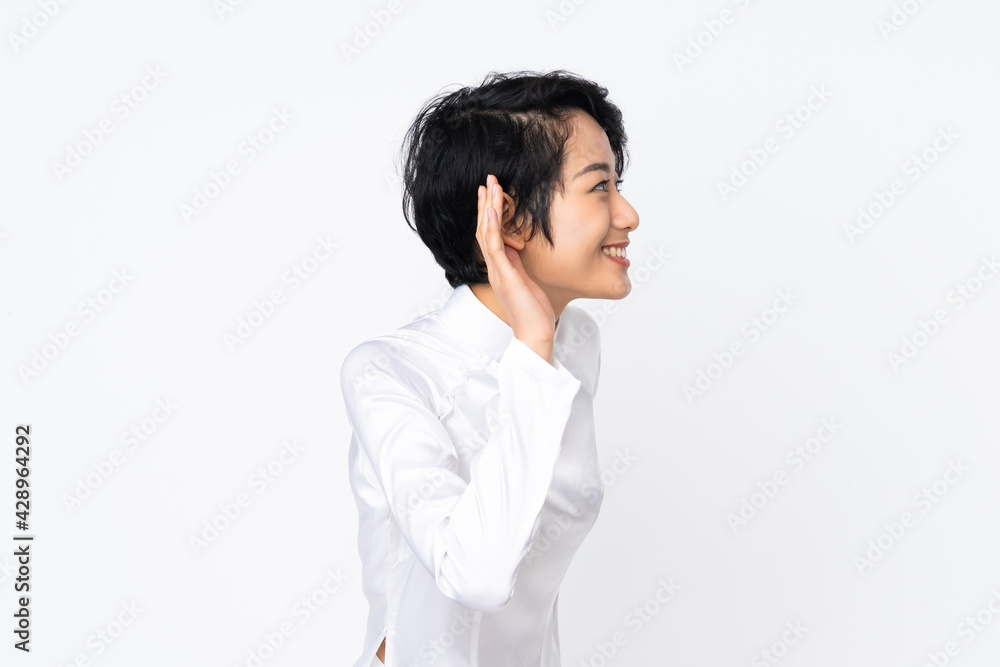 Young Vietnamese woman with short hair wearing a traditional dress over isolated white background listening to something by putting hand on the ear Young Vietnamese woman with short hair wearing a traditional dress over isolated white background listening to something by putting hand on the ear