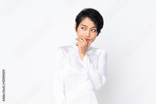 Young Vietnamese woman with short hair wearing a traditional dress over isolated white background having doubts and with confuse face expression