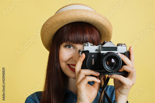 Close up of beautiful young woman photographer in straw hat taking a picture and looking in viewfinder on retro digital mirrorless photo camera, posing on studio isolated yellow background.