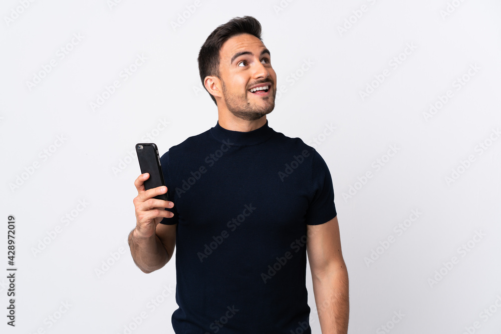 Young caucasian man using mobile phone isolated on white background looking up while smiling