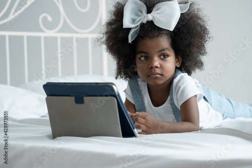 Little cute African kid lying down on bed absent minded and looking other way while using tablet computer to learn online class at home photo