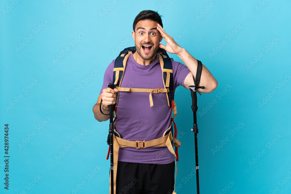 Young caucasian man with backpack and trekking poles isolated on blue background with surprise expression Young caucasian man with backpack and trekking poles isolated on blue background with surprise expression