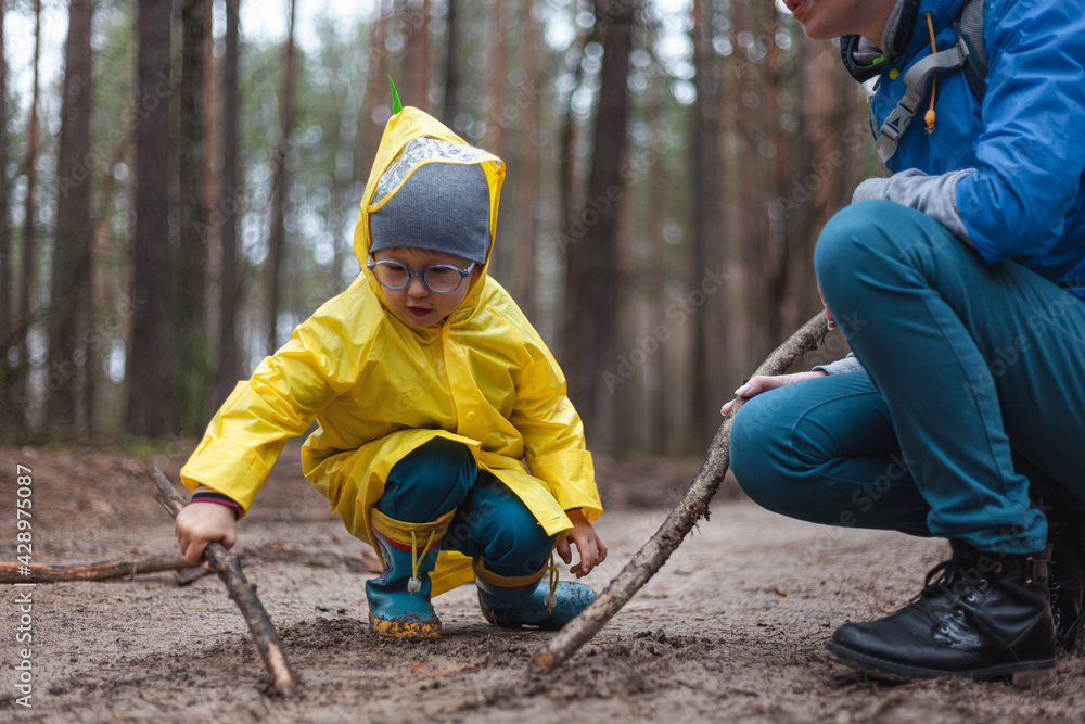 Mom and child walking along the forest road after rain in raincoats, together draw on the sand with wooden sticks Mom and child walking along the forest road after rain in raincoats, together draw on the sand with wooden sticks