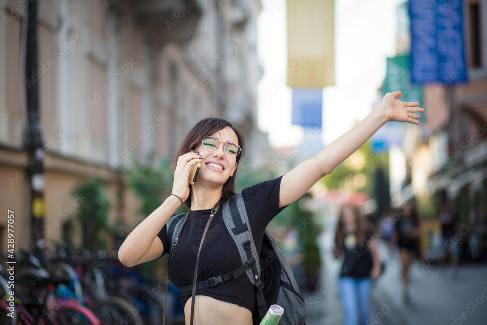 Smiling young woman on street talking on smart phone and waving.