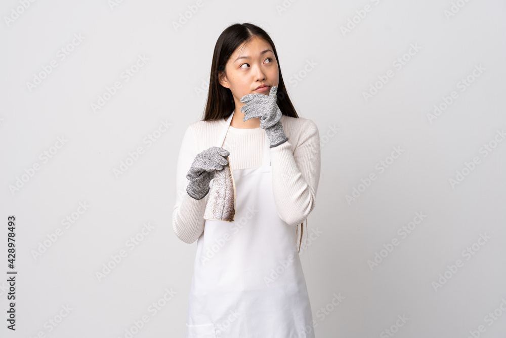 Chinese fishmonger wearing an apron and holding a raw fish over isolated white background having doubts and with confuse face expression Chinese fishmonger wearing an apron and holding a raw fish over isolated white background having doubts and with confuse face expression