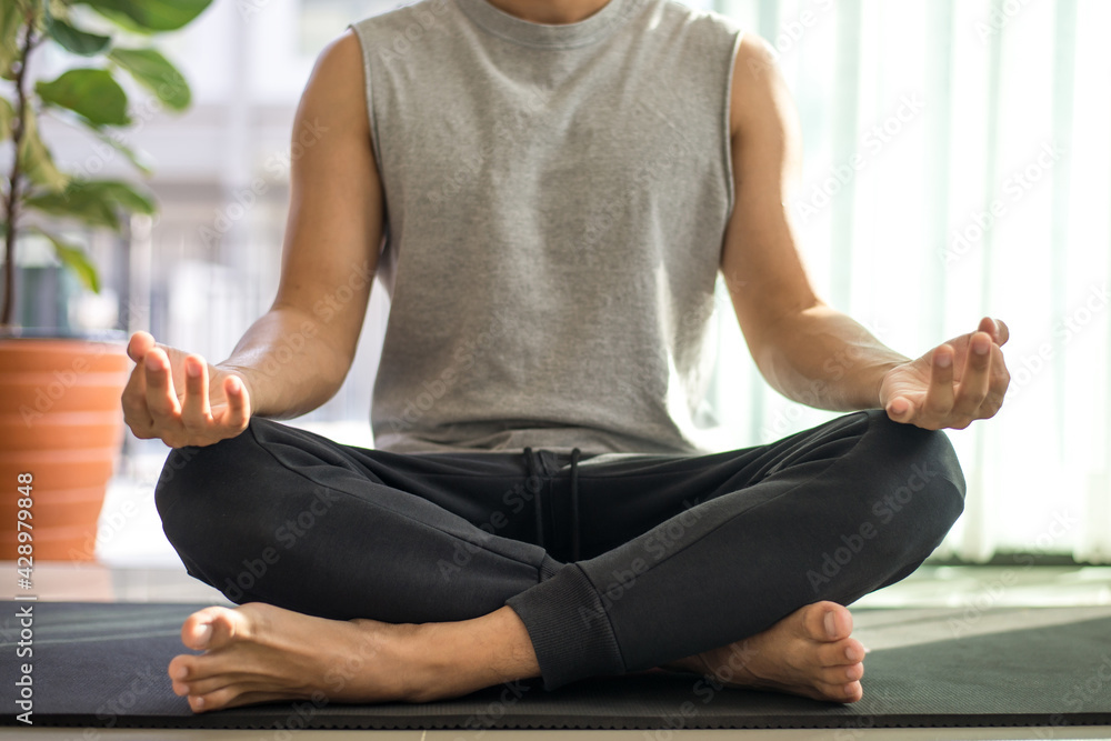 Man in a gray sleeveless shirt and black pants sat on the exercise mat practicing meditation, sitting in a lotus pose alone indoors at home. During the quarantine due to the spread of the coronavirus.