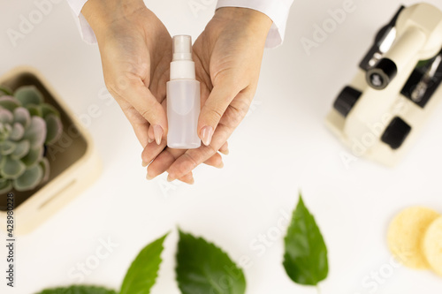 cosmetologist female hands hold plastic bottle of cosmetic lotion or serum on white background of cosmetology lab with microscope. Natural herbal cosmetics advertisement