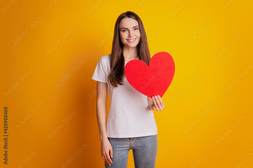 Photo of dreamy lady hold paper heart figure wear white t-shirt posing on yellow background Photo of dreamy lady hold paper heart figure wear white t-shirt posing on yellow background