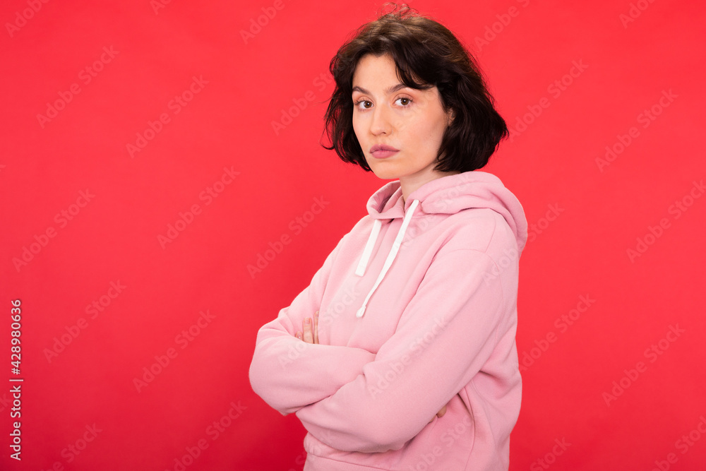Young brunette woman in trendy hipster hoodie posing with crossed arms on isotaled red background. Concept of confident strong female person student, feminism, fight for women's rights. Copy space Young brunette woman in trendy hipster hoodie posing with crossed arms on isotaled red background. Concept of confident strong female person student, feminism, fight for women's rights. Copy space