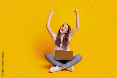 Photo of cheerful lady hold netbook look up empty space celebrate win wear white t-shirt posing on yellow background © Tetiana