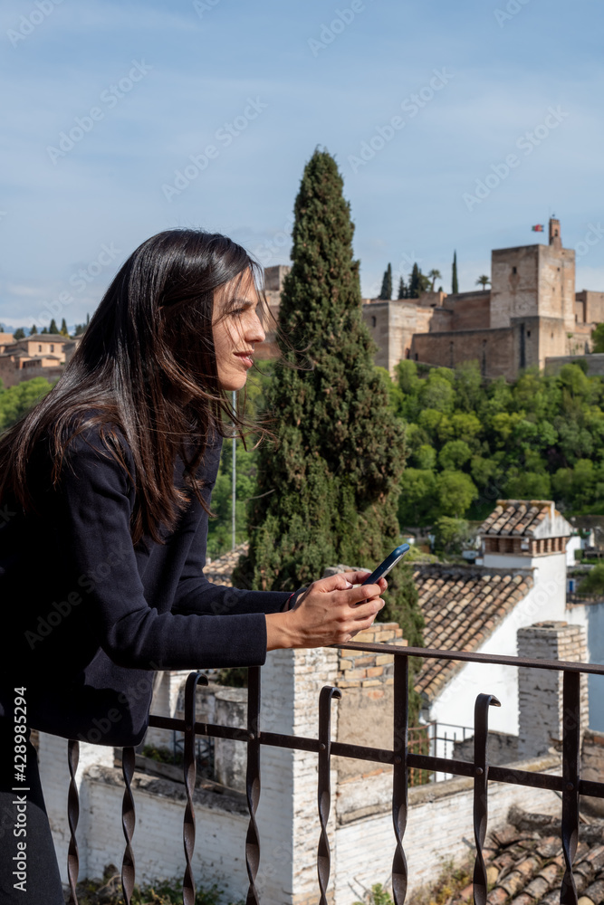 Hispanic female tourist with phone standing on terrace with views to Alhambra fortress in Granada, Spain