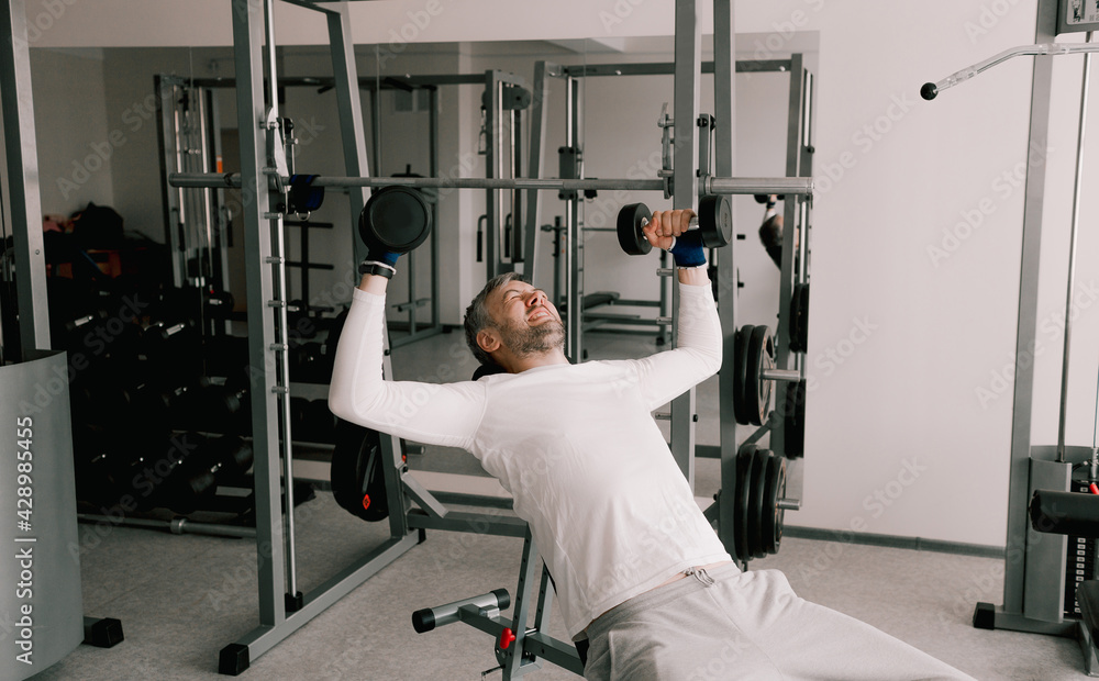 Portrait of a young man doing physical exercises, training with dumbbells sitting on a bench in the gym. In a white T-shirt. Portrait of a young man doing physical exercises, training with dumbbells sitting on a bench in the gym. In a white T-shirt.