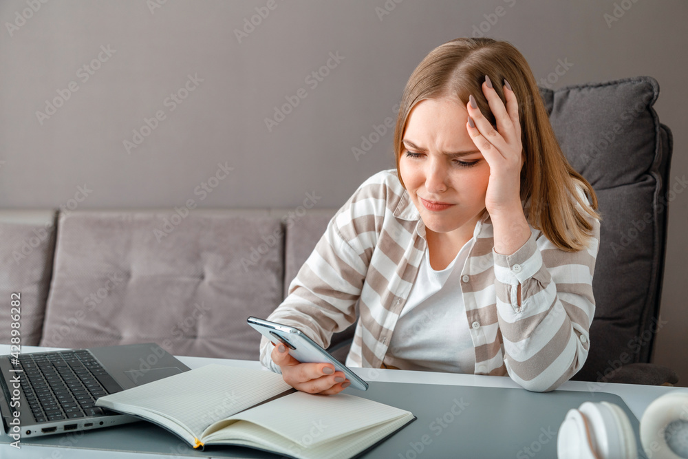 Young woman upset under stress. Confused woman holding head received bad news via smartphone at work. Teen girl looks at smartphone screen during Online work or education negative emotions depression. Young woman upset under stress. Confused woman holding head received bad news via smartphone at work. Teen girl looks at smartphone screen during Online work or education negative emotions depression.