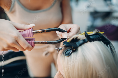 Hairdresser female making hair extensions to young woman with blonde hair in beauty salon