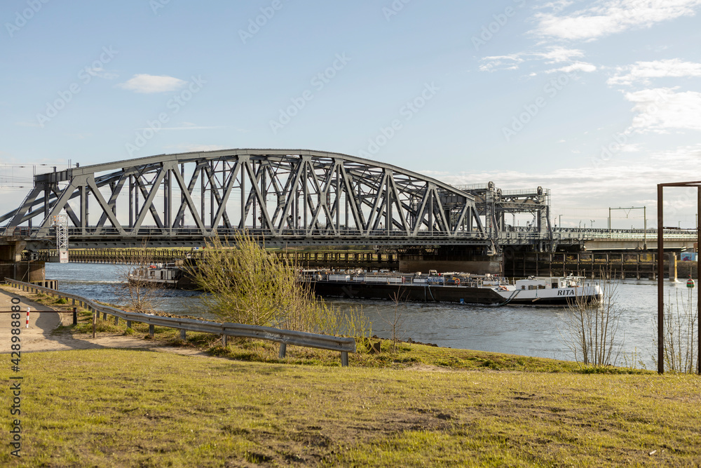 Steel train and traffic draw bridge over the river IJssel with a cargo ship passing by near the Dutch Hanseatic city of Zutphen, The Netherlands, seen from the floodplains against a blue sky Steel train and traffic draw bridge over the river IJssel with a cargo ship passing by near the Dutch Hanseatic city of Zutphen, The Netherlands, seen from the floodplains against a blue sky