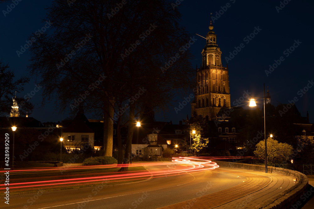 Light streaks of passing cars on meandering town road lit up by city lights with Walburgiskerk cathedral towering behind into the night. Long exposure blue hour cityscape.