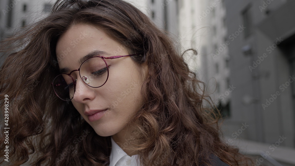 Portrait Of Young Girl On City Street. Pretty Girl With Curly Long Hair And Glasses. She Smiles. Portrait Of Young Girl On City Street. Pretty Girl With Curly Long Hair And Glasses. She Smiles.
