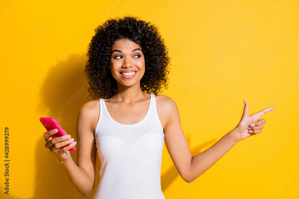 Photo of attractive dark skin brunette girl wear white tank-top look point empty space hold phone isolated on yellow color background Photo of attractive dark skin brunette girl wear white tank-top look point empty space hold phone isolated on yellow color background