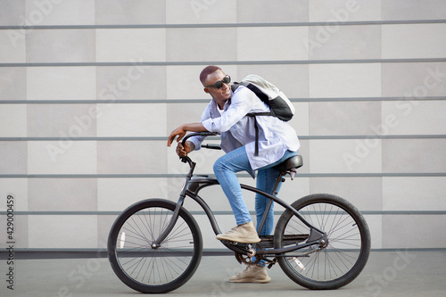 Fototapeta Naklejka Na Ścianę i Meble -  Portrait of handsome black guy with backpack and sunglasses riding bicycle near brick wall outside