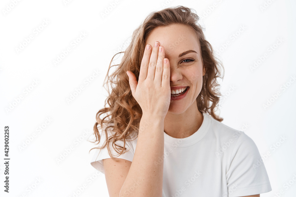 Candid attractive woman hide half of face behind hand and smile, laughing and showing natural happy face expression, standing over white background Candid attractive woman hide half of face behind hand and smile, laughing and showing natural happy face expression, standing over white background