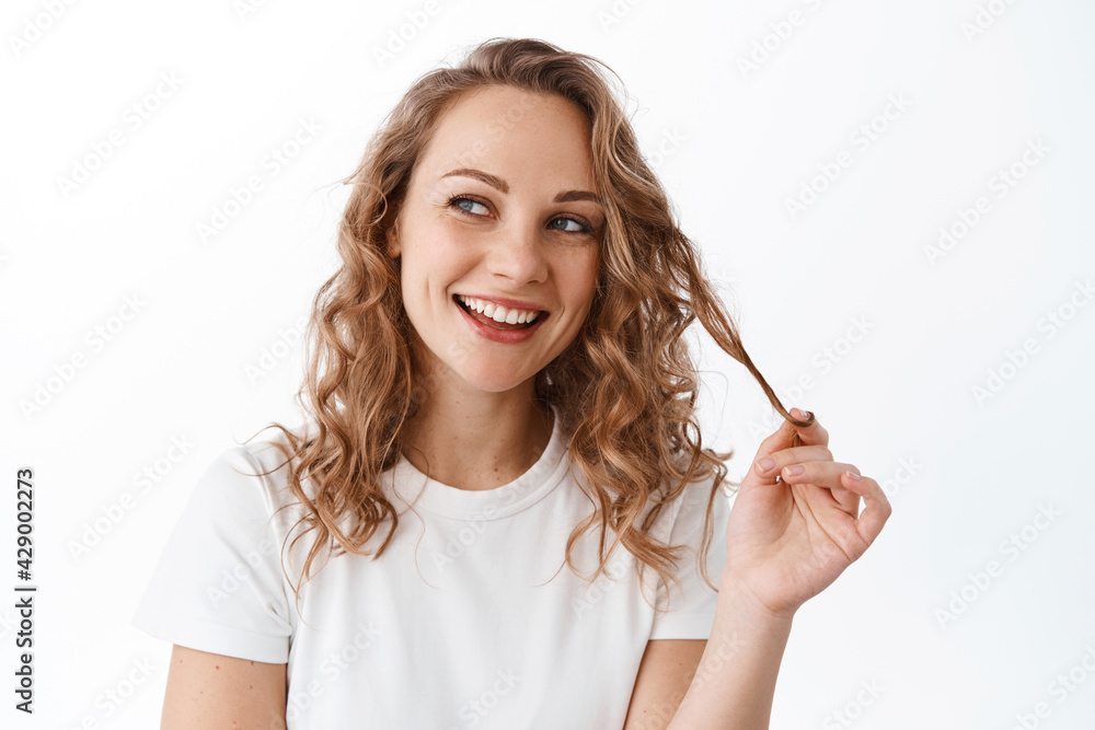 Cute blond curly girl smiling, looking thoughtful aside, playing with hair strand and staring at promotional text copy space, standing over white background Cute blond curly girl smiling, looking thoughtful aside, playing with hair strand and staring at promotional text copy space, standing over white background