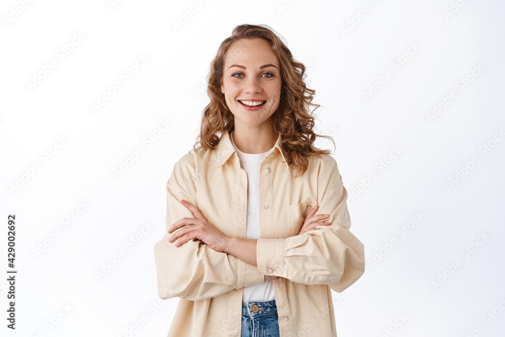 Young confident woman with blond curly hair, cross arms on chest and look determined, smiling self-assured, standing over white background Young confident woman with blond curly hair, cross arms on chest and look determined, smiling self-assured, standing over white background