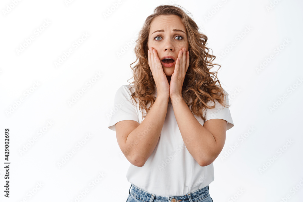Shocked young woman in panic, holding hands on face, screaming and looking concerned, express pity and nervous emotions, standing over white background Shocked young woman in panic, holding hands on face, screaming and looking concerned, express pity and nervous emotions, standing over white background