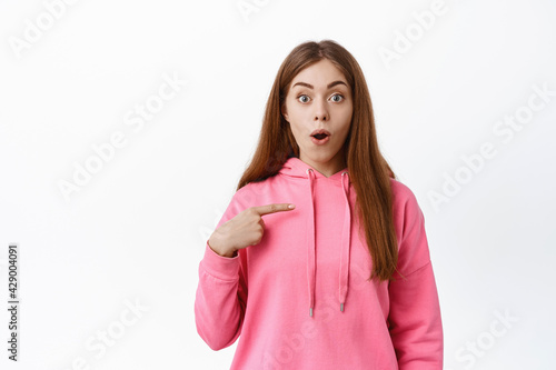 Image of girl points at herself, gasps and looks surprised, being chosen, picked to participate, stare amazed at camera, stands against white background