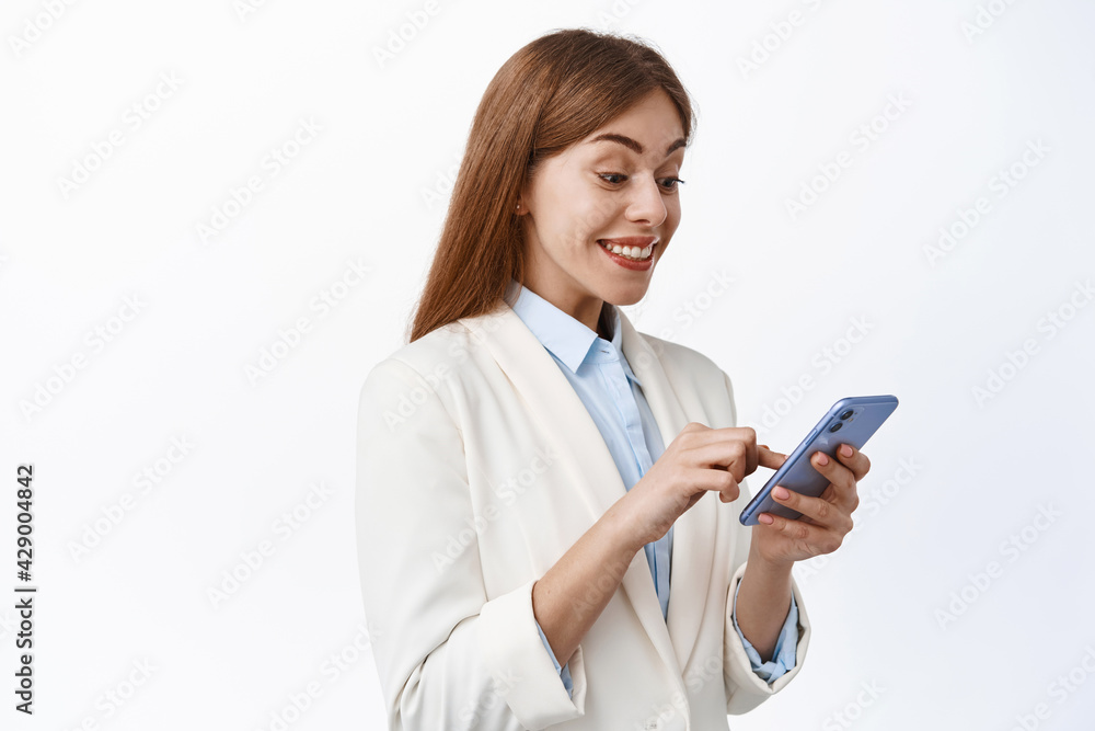 Happy female ceo manager in business suit, looks at smartphone screen and smile pleased, reading message on phone, standing over white background Happy female ceo manager in business suit, looks at smartphone screen and smile pleased, reading message on phone, standing over white background