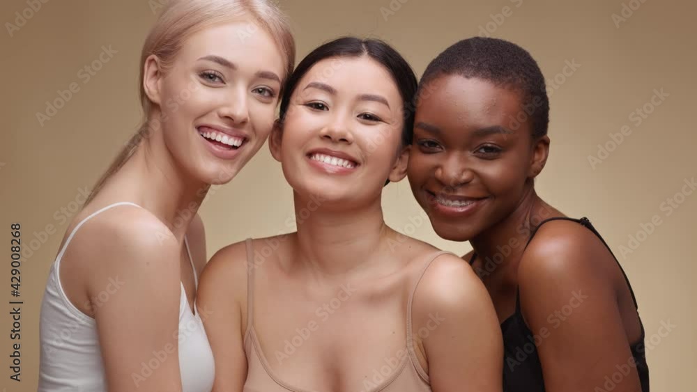Studio portrait of three happy multiethnic ladies laughing to camera, enjoying friendship, hugging over beige background Studio portrait of three happy multiethnic ladies laughing to camera, enjoying friendship, hugging over beige background