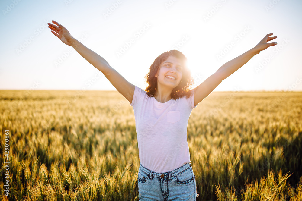 A smiling girl stands against the background of a field with wheat with divorced hands. Photo with a woman in a pink T-shirt on a sunset background. A smiling girl stands against the background of a field with wheat with divorced hands. Photo with a woman in a pink T-shirt on a sunset background.