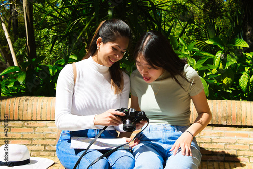 Two Asian women on holiday. They are sitting in a park looking at the pictures on the camera. On her legs she is holding a tourist map of the city. Two young women are happy looking at the pictures.