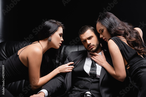 Foto successful businessman near passionate women seducing him on leather couch on black