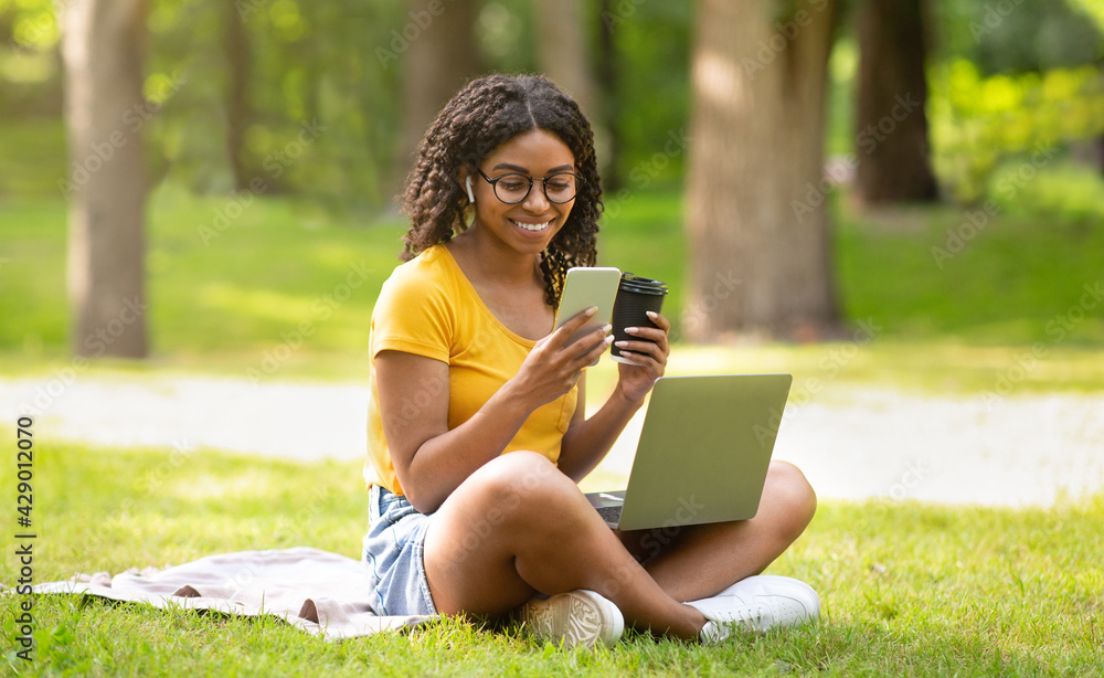 Funky black girl with takeout coffee, mobile phone and laptop studying online at park Funky black girl with takeout coffee, mobile phone and laptop studying online at park