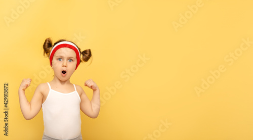 Excited fitness child posing in sportswear,red wristbands,headband over yellow studio background. Children sport,advertising,shopping concept.Gym workout.Childhood activity.Sport.Health and energy