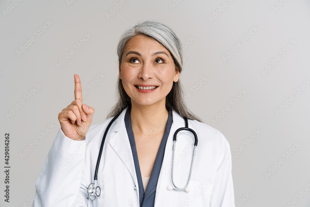 Asian mature woman in medical uniform smiling and pointing finger upward Asian mature woman in medical uniform smiling and pointing finger upward