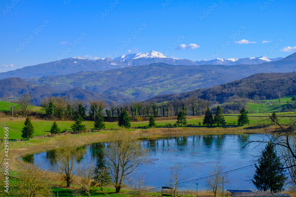Beautiful spring landscape with mountain lake and mountains covered with snow across blue sky. Fir trees, and trees reflected in water.