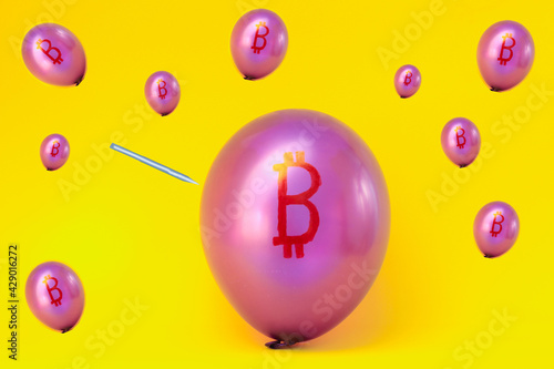 A pattern of balloons depicting pink bitcoin on a yellow background with a sharp object piercing the balloon. The concept of rapid decline of cryptocurrency.