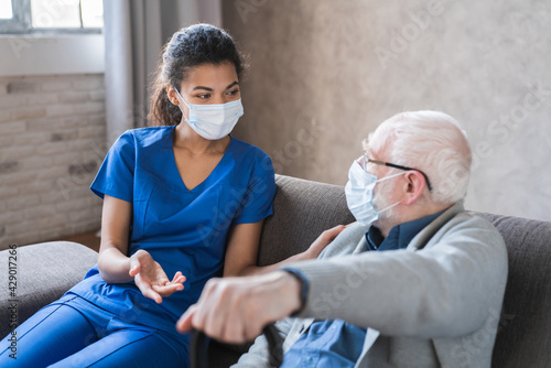 Doctor or female caregiver with senior man grandfather wearing protective mask to protect from Covid 19 at home or nursing home. Doctor talking consulting elderly patient at home photo
