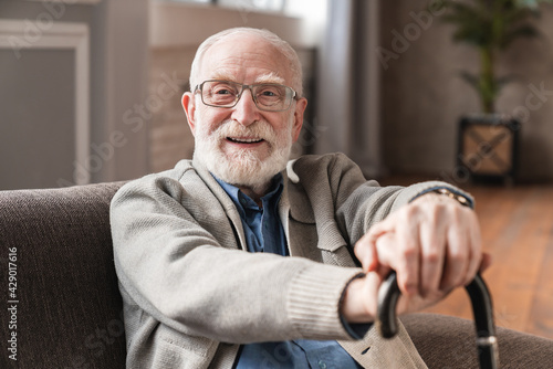 Close up photo of grey haired grandparent with walking stick head lean on hand wearing casual outfit sitting on cozy sofa. Physically disabled positive old grandfather seated on couch resting at home.