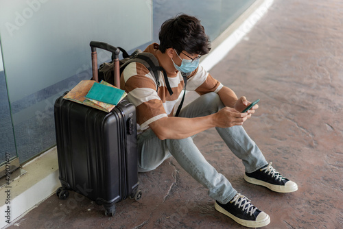 Travel. A man wearing glasses with a striped shirt and backpack, wear mask, sit on the floor. using a phone during travel with travel stuffs on the suitcase which on right side photo