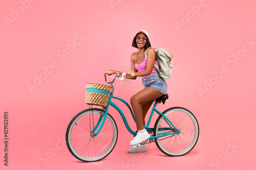 Full length portrait of lovely black woman in casual summer clothes riding bicycle over pink studio background © Prostock-studio