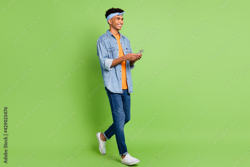 Photo of guy hold smartphone look empty walk wear headband jeans shirt sneakers isolated green color background Photo of guy hold smartphone look empty walk wear headband jeans shirt sneakers isolated green color background
