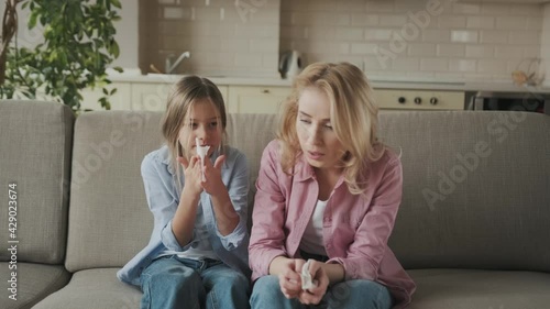 Sad caucasian mother and daughter with a cold are sitting at home on the sofa with napkins, sneezing, coughing, runny nose. Cold, flu, illness
