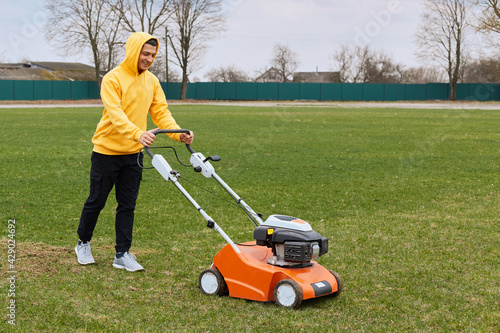 Happy smiling handsome man wearing yellow hoodie and black pants, trimming grass on field with grass-cutter, worker mow lawn with lawn mower, expressing positive emotions.