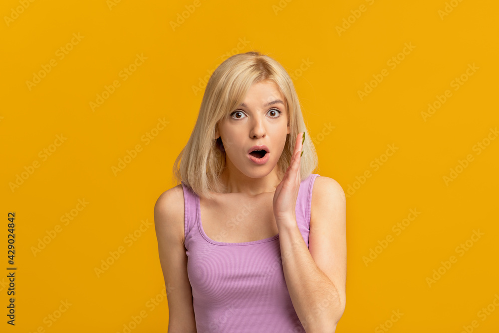 Shocking gossip. Emotional blonde woman keeping hand near opened mouth telling secret over yellow studio background Shocking gossip. Emotional blonde woman keeping hand near opened mouth telling secret over yellow studio background