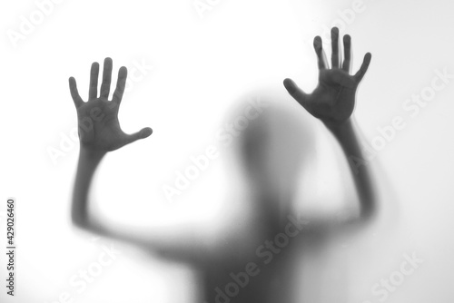 Silhouette scared boy stand behind glass door. Kid showing two hands.