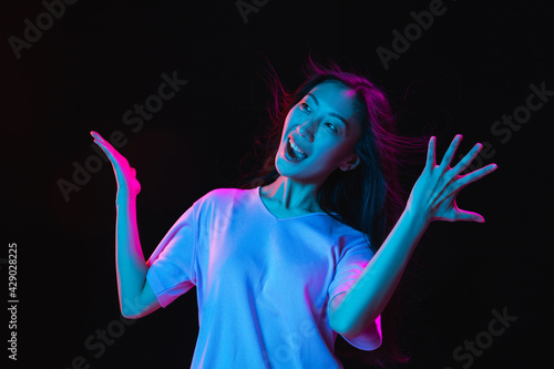 Asian young woman's portrait on dark studio background in neon. Concept of human emotions, facial expression, youth, sales, ad.