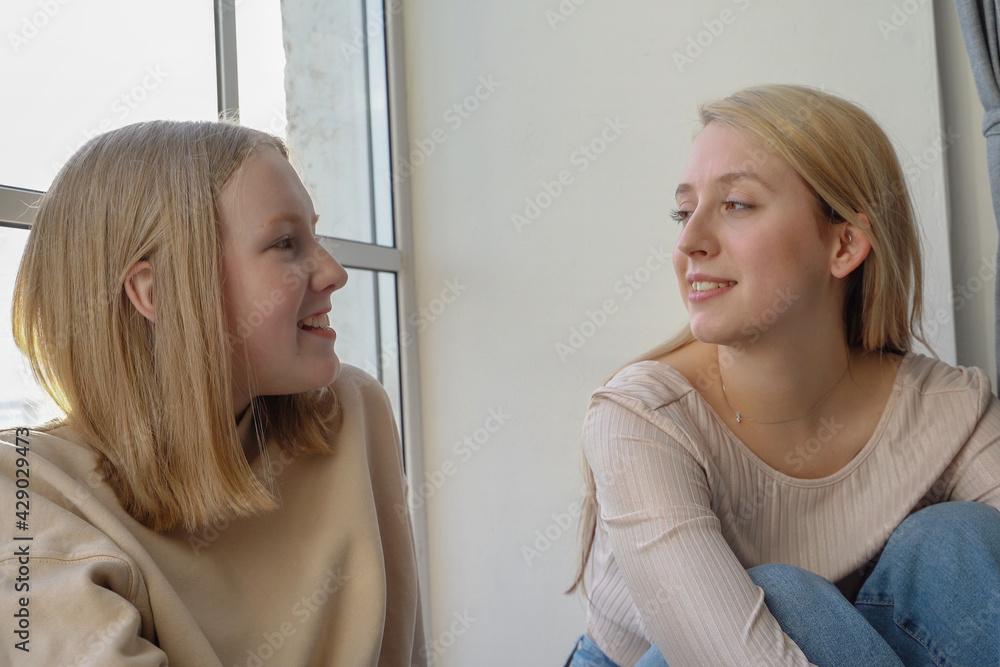 Two sisters sitting near the window and talk about some news. Surprised and smiled girl face. Chatting.